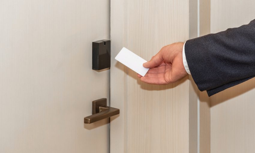A person holds a white card key close to a hotel room door lock. The door is light-colored and made of wood.
